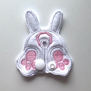 May include: White fabric bunny-shaped baby bib with pink embroidered paw prints and a pink nose. The bib has two white snap buttons.