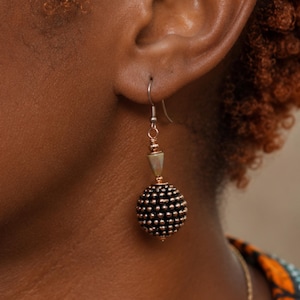 May include: A drop earring featuring a large, spherical black bead with a textured, studded surface. The bead is accented with a small, conical, iridescent stone bead above it, connected by rose gold-toned metal components to a silver-toned hook.