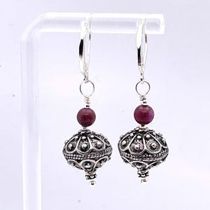 May include: A pair of silver earrings with a lever back closure. Each earring features a small, round, dark red bead above a detailed, silver, orb-shaped pendant. The pendants have an ornate, textured design.