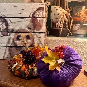 May include: Two decorative fabric pumpkins, one with a floral pattern and the other a solid purple, with fall leaves and berries. The pumpkins are sitting on a wooden surface in front of a framed painting of a pig.