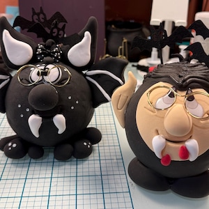 May include: Two round clay figurines featuring vampire themes. The left figure is black with white wings, fangs, and glasses. The right figure has a peach-coloured face, black hair, glasses, and fangs with red drops. Both have black bat toppers.