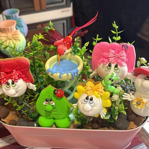 May include: A decorative arrangement in a pink metal container. It features whimsical, cartoon-like figures, including a red bird, leaf, and flower shapes, with faces and bows. Green foliage and a blue water dish add to the scene.