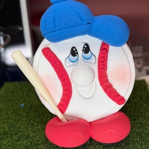 K&ouml;nnte beinhalten: Eine skurrile Baseball-Figur. Der wei&szlig;e Baseball hat rote N&auml;hte, eine blaue Kappe und rote F&uuml;&szlig;e. Er h&auml;lt einen beigen Baseballschl&auml;ger und hat einen traurigen Gesichtsausdruck. Die Figur steht auf einer gr&uuml;nen, grasbewachsenen Oberfl&auml;che.