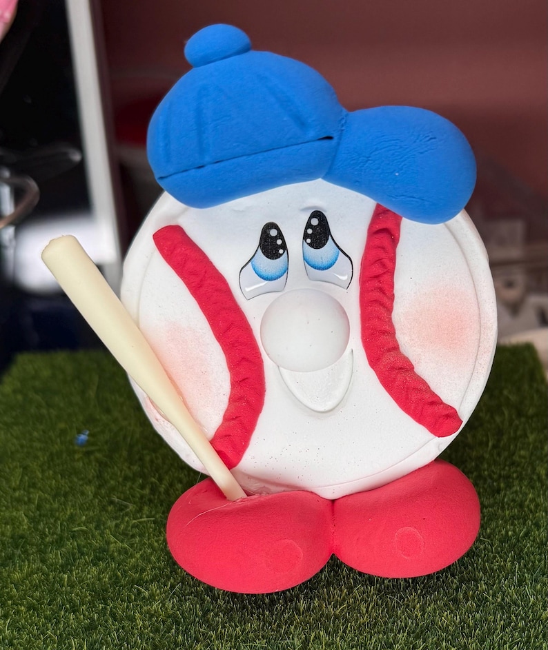 K&ouml;nnte beinhalten: Eine skurrile Baseball-Figur. Der wei&szlig;e Baseball hat rote N&auml;hte, eine blaue Kappe und rote F&uuml;&szlig;e. Er h&auml;lt einen beigen Baseballschl&auml;ger und hat einen traurigen Gesichtsausdruck. Die Figur steht auf einer gr&uuml;nen, grasbewachsenen Oberfl&auml;che.