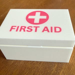 May include: A white first aid box with a red cross symbol and the words "FIRST AID" printed on the lid. The box is rectangular and appears to be made of a solid material.