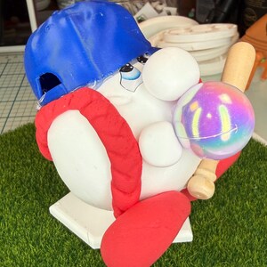 K&ouml;nnte beinhalten: Eine skurrile Baseball-Figur. Sie zeigt eine wei&szlig;e Figur mit einer blauen Baseballkappe und einer roten Uniform. Die Figur h&auml;lt einen Baseball und einen Holzschl&auml;ger. Der Sockel ist wei&szlig; und hat die Form der Homeplate.