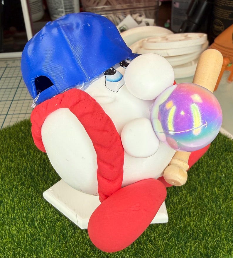 K&ouml;nnte beinhalten: Eine skurrile Baseball-Figur. Sie zeigt eine wei&szlig;e Figur mit einer blauen Baseballkappe und einer roten Uniform. Die Figur h&auml;lt einen Baseball und einen Holzschl&auml;ger. Der Sockel ist wei&szlig; und hat die Form der Homeplate.