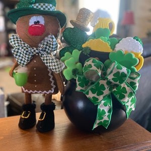 May include: A gingerbread man wearing a green and white checkered hat and a green bow tie holds a green cup.  A black cauldron is filled with green shamrock cookies, a green and white ribbon, and a gold top hat.