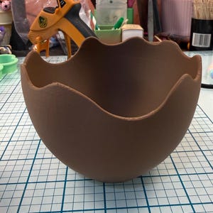 May include: A brown, egg-shaped bowl with a scalloped rim. The bowl is made of a solid material and sits on a white grid surface. The bowl's interior is empty, and the exterior has a matte finish.