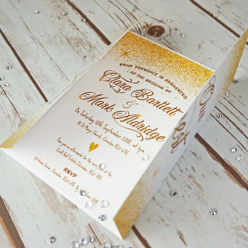 Puede incluir: Invitaci&oacute;n de boda con purpurina dorada sobre cartulina blanca. La invitaci&oacute;n dice "Se solicita su presencia en la boda de Clara Bartlett & Mark Aldridge el s&aacute;bado 10 de septiembre de 2018 a la 1:00 pm en la Iglesia de St. Mary's, Clevedon BS21 GAU."