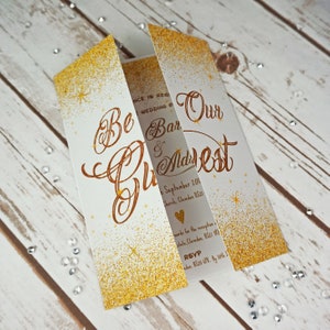 Puede incluir: Invitaci&oacute;n de boda con purpurina dorada y el texto "Be Our Guest" en letra cursiva. La invitaci&oacute;n incluye la fecha, la iglesia y la informaci&oacute;n de RSVP.