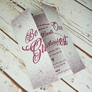 Puede incluir: Una invitaci&oacute;n de boda blanca con purpurina rosa y el texto "Be Our Guest" y "Bart & Madrid Cat" con la fecha "Septiembre de 2018, una iglesia, Claveden BS21 6AU".