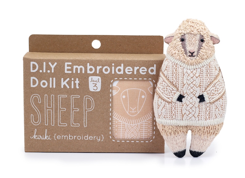May include: A brown cardboard box with a white label that says "D.I.Y Embroidered Doll Kit Level 3 SHEEP kiriki (embroidery)". The box contains a kit to make a sheep doll. A finished sheep doll is shown next to the box. The sheep doll is wearing a white sweater and has a brown face.