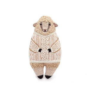 May include: A plush sheep toy wearing a white knitted sweater with a cable knit pattern. The sheep has a brown face and black hooves.