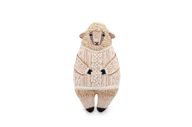 May include: A plush sheep toy wearing a white knitted sweater with a cable knit pattern. The sheep has a brown face and black hooves.
