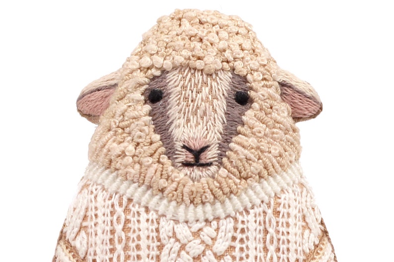 May include: Close-up of a white sheep with a brown and pink face, wearing a white knitted sweater with a cable knit pattern.