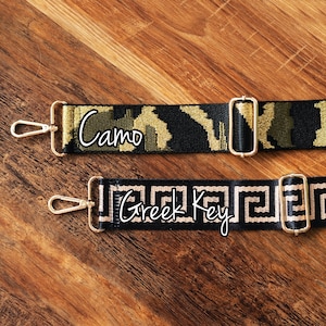 May include: Two black and gold adjustable straps with gold clasps. The top strap has a camouflage pattern and the text "Camo". The bottom strap has a Greek key pattern and the text "Greek Key".