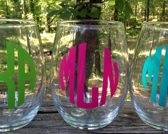May include: Three clear glass wine glasses with personalized monograms. The monograms are in green, pink, and teal.