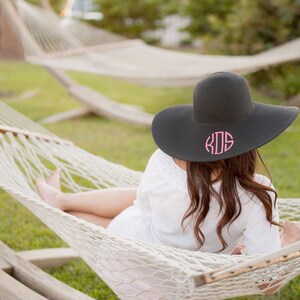 May include: A woman wearing a black sun hat with pink monogrammed initials "KDS" is relaxing in a white hammock.