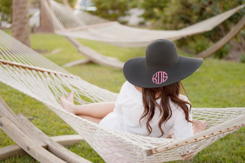May include: A woman wearing a black sun hat with pink monogrammed initials "KDS" is relaxing in a white hammock.