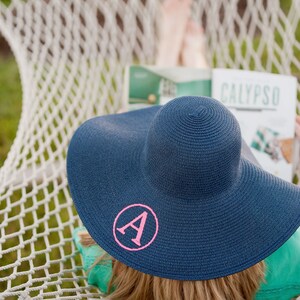 May include: A woman relaxes in a hammock wearing a blue sun hat with a pink letter A embroidered on the front. The hat is wide-brimmed and has a woven texture. The woman is reading a magazine with the word "CALYPSO" printed on the cover.