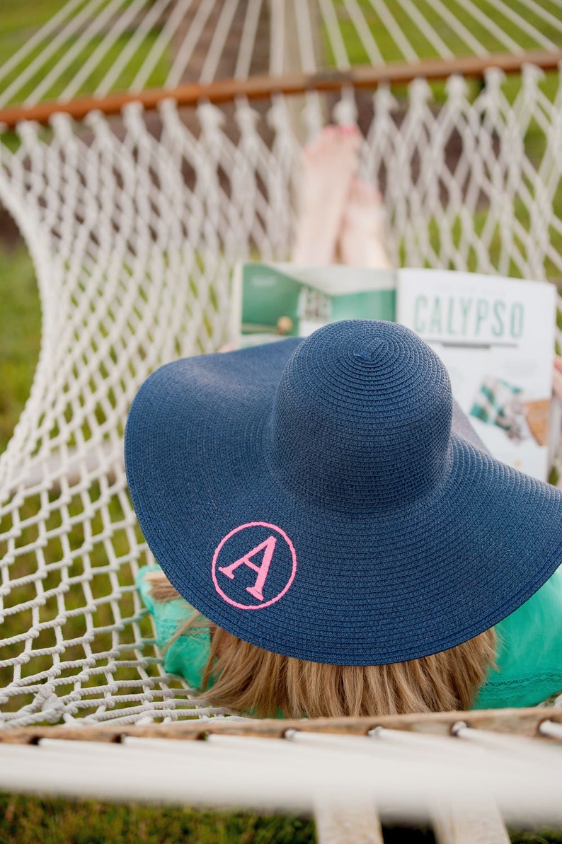 May include: A woman relaxes in a hammock wearing a blue sun hat with a pink letter A embroidered on the front. The hat is wide-brimmed and has a woven texture. The woman is reading a magazine with the word "CALYPSO" printed on the cover.