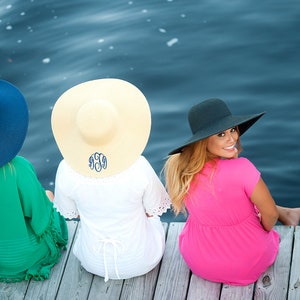 May include: Three women wearing straw and fabric sun hats with monograms on a wooden dock by a body of water.
