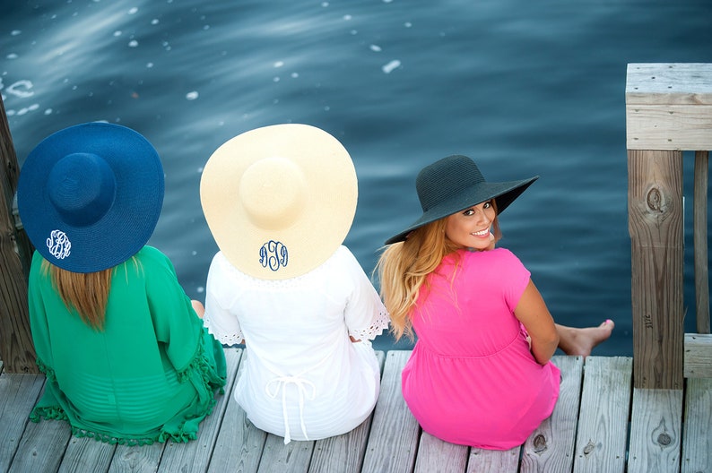 May include: Three women wearing straw and fabric sun hats with monograms on a wooden dock by a body of water.