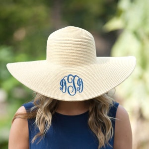 May include: A wide-brimmed straw sun hat with a blue embroidered monogram. The hat is beige and has a wide, floppy brim.