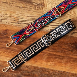 May include: Two colorful woven straps with gold metal clasps. The strap on the left has a geometric pattern in red, blue, yellow, and black with the word "Aztec" written on it. The strap on the right has a black and gold pattern with the words "Gold Greek Key" written on it.