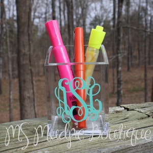 May include: A clear acrylic desk organizer with a teal monogram "S" on the front. The organizer holds three markers: a pink marker, an orange marker, and a yellow highlighter.