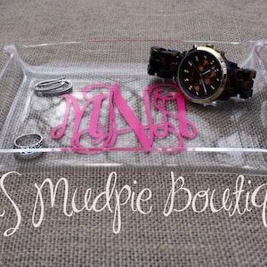 May include: A clear acrylic tray with a pink monogram "M.N.A." and a gold watch with a brown face and a brown leather band. The tray also contains two silver rings. The tray is on a brown burlap background.  MS Mudpie Boutique.