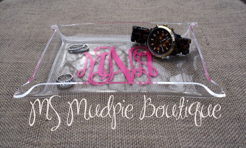 May include: A clear acrylic tray with a pink monogram "M.N.A." and a gold watch with a brown face and a brown leather band. The tray also contains two silver rings. The tray is on a brown burlap background.  MS Mudpie Boutique.