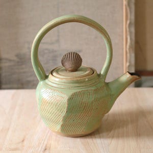 May include: A light green ceramic teapot featuring a high arched handle, a spout, and a lid topped with a small, textured seashell-shaped knob. The body of the teapot displays a series of vertical, incised line patterns across its rounded surface.