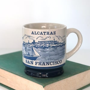 May include: A ceramic mug with a beige speckled finish, featuring a blue illustration of Alcatraz Island and a sailboat. The words "ALCATRAZ" and "SAN FRANCISCO" are printed on the mug. The mug has a dark blue base and a curved handle.