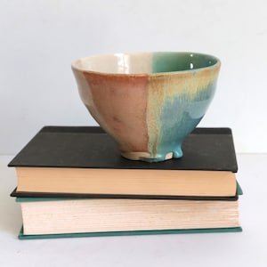 May include: A handmade ceramic bowl with a unique color scheme. The bowl is divided into sections of beige, brown, and teal. It sits atop two stacked books with black covers and tan pages. The bowl's design is asymmetrical and organic.