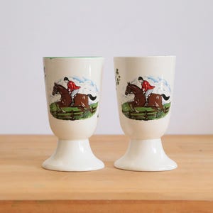 May include: Two white ceramic cups with a green rim and a horse racing scene printed on the side. The cups have a pedestal base.