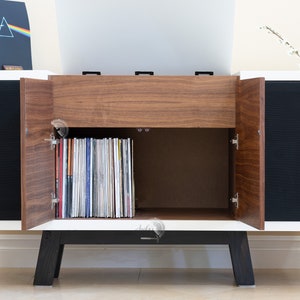 May include: A white and walnut record cabinet with two doors open revealing a collection of vinyl records. The cabinet has black legs and a black speaker grille on each side.