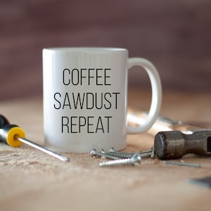 May include: White ceramic coffee mug with the text "Coffee Sawdust Repeat" in black lettering. The mug is sitting on a wooden surface with a screwdriver, screws, and a hammer.