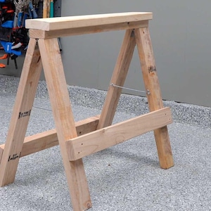 May include: A wooden sawhorse with a rectangular top and angled legs. The sawhorse is constructed from light-colored lumber and features a horizontal support beam near the base. A metal wire provides additional stability.