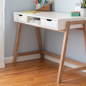 May include: White desk with two drawers and a shelf, made of wood with a light wood finish. The desk has a simple, modern design and is perfect for a home office or bedroom.