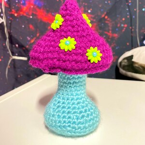 Könnte beinhalten: Ein gehäkelter Pilz mit einem rosa Hut und einem hellblauen Stiel. Der Hut ist mit fünf gelben Blüten mit blauen Zentren verziert.