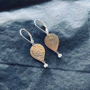 Op de afbeelding: Een paar gouden oorringen in de vorm van een traan met een zilveren haak en een helder kristal dat aan elke oorring hangt.