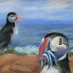 May include: Two puffins on a rocky shore with blue water in the background. One puffin is standing on a rock and the other is holding a beak full of fish.