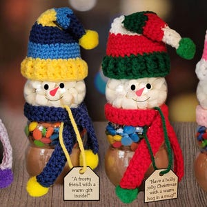 May include: Four snowman candy jars with crochet hats and scarves in purple, blue, red, and pink. Each jar has a white marshmallow body, a candy-filled base, and a tag. The image includes the text "Crochet Hat and Scarf for a Snowman Candy Jar!"