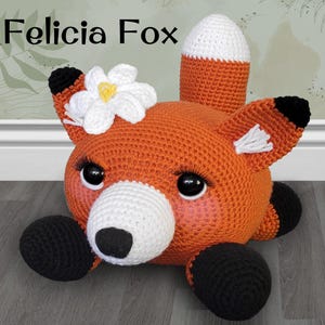 May include: A crocheted orange fox with black eyes, a white nose, and black paws. The fox is wearing a white flower with a yellow center on its head. The fox is lying on its belly with its head turned to the side. The text "Felicia Fox" is written above the fox.