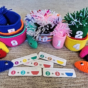 May include: A colorful crochet math learning set featuring nesting bowls with numbers, crochet fish, and cards with addition problems. The bowls are in various colors, and the fish are red, yellow, blue, orange, and purple.