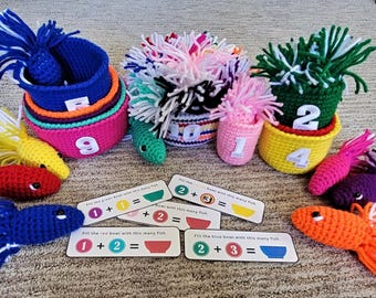 Puede incluir: Un colorido juego de aprendizaje de matemáticas de ganchillo con cuencos apilables numerados, peces de ganchillo y tarjetas de suma. Los peces son rojos, amarillos, azules, naranjas y morados. Las tarjetas muestran problemas de suma.