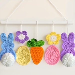 Op de afbeelding: Een kleurrijke gehaakte paasversiering met twee konijnen, twee eieren, twee bloemen en een wortel. De versiering hangt aan een witte houten stok.