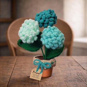 Puede incluir: Un arreglo floral hecho a mano con ganchillo en una pequeña maceta de terracota. El arreglo presenta tres flores de hortensia en tonos de verde menta y turquesa, con hojas verdes. Una etiqueta atada con un lazo turquesa dice "Forever, blooming, just like your heart!"